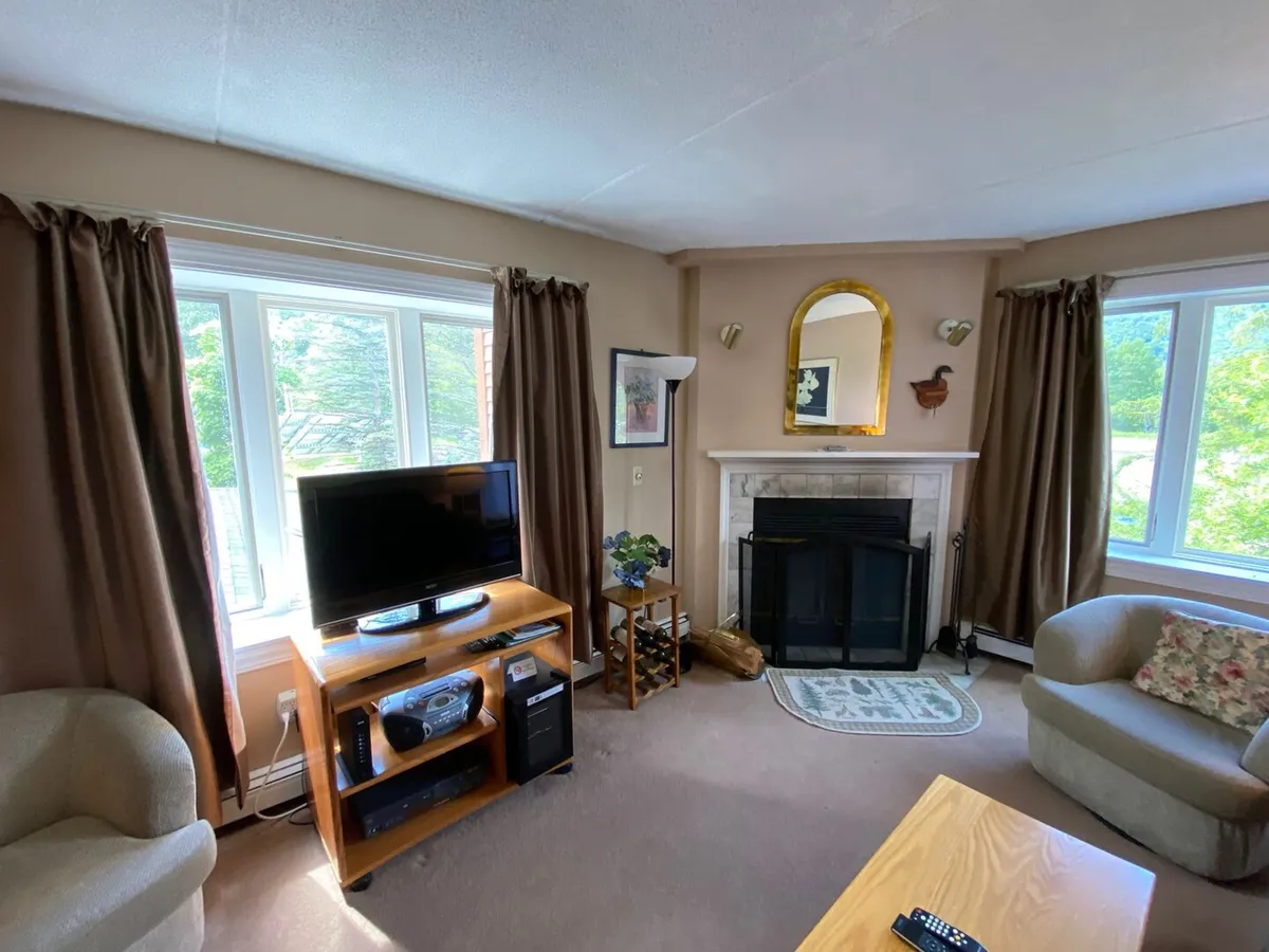 This inviting living room features a sofa, fireplace, large windows, and a television. Abundant natural light streams in through the expansive windows, while the seating area is oriented for both relaxation and socializing. The view through the windows hints at the possibility of mountain scenery and the adjacent balcony.