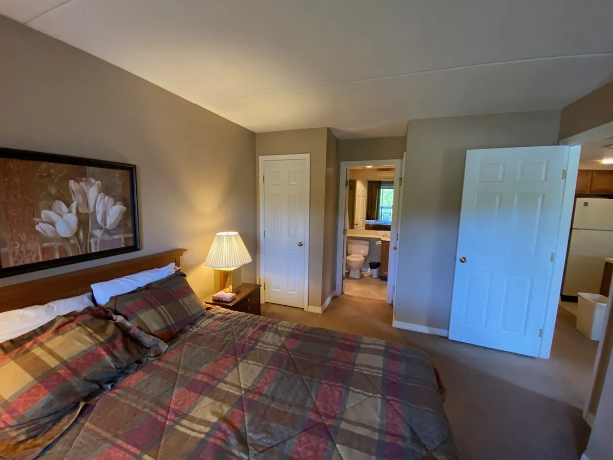 A cozy bedroom with plaid bedding, soft morning light filtering through the window, a bedside table with a lamp, and subtle floral artwork on the wall. The doorway gives a glimpse into the kitchen space, suggesting proximity to morning routines.