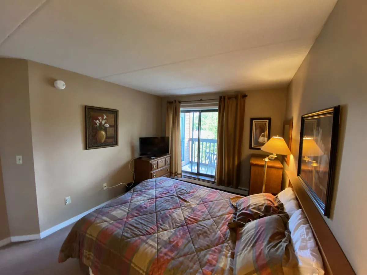A bright and inviting bedroom scene, this image highlights a comfortable bed positioned next to a window with a view. Sunlight streams in, and additional features like a lamp and television offer extra comfort and relaxation options.