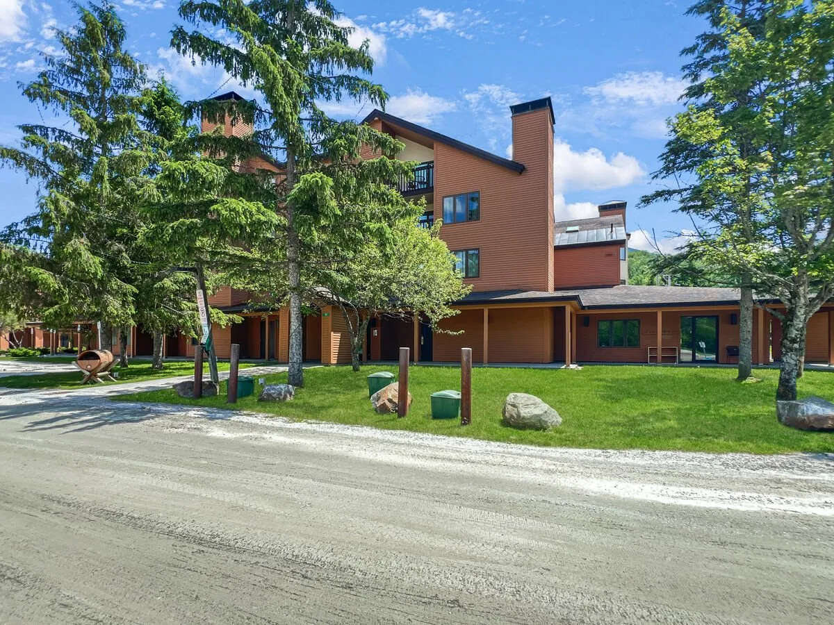 This image features the exterior facade of the building set among trees and a landscaped gravel path. The welcoming scene includes a porch area that hints at a communal, relaxed atmosphere, inviting guests to imagine stepping directly outside and enjoying immediate access to the outdoors.